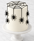 A white frosted cake decorated with black spider and web designs, displayed on a white marble cake stand against a light marble surface by The Makery Cake Co in Denver.