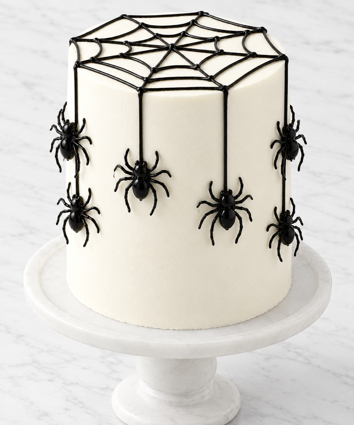 A white frosted cake decorated with black spider and web designs, displayed on a white marble cake stand against a light marble surface by The Makery Cake Co in Denver.