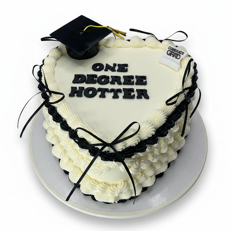 Heart-shaped cake with a graduation cap and 'ONE DEGREE HOTTER' text on a wooden surface.
