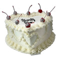 White heart-shaped cake with vintage piping snd cherries on a white stand against a light wooden panel background