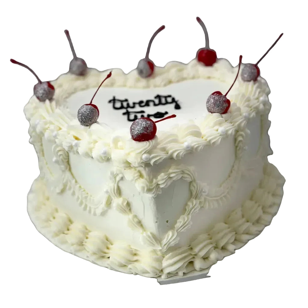 White heart-shaped cake with vintage piping snd cherries on a white stand against a light wooden panel background